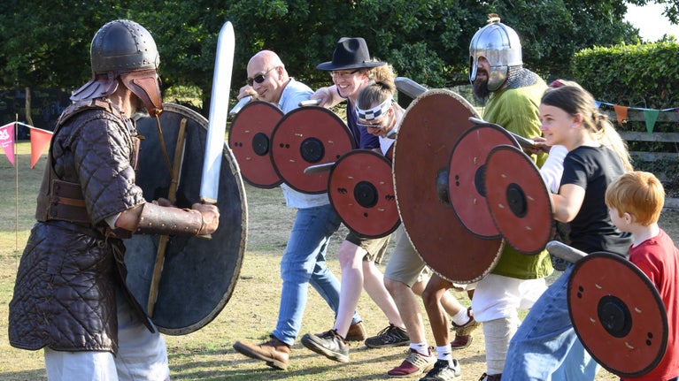 A row of children and adults line up with red circular shields to train like a warrior in the grounds at Sutton Hoo